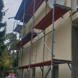 Ariel Kamiński - Elewacja budynku w trakcie malowania, widoczne rusztowania, pomalowana górna część ściany na beżowo, dolna część w szarym podkładzie, antena satelitarna na ścianie.