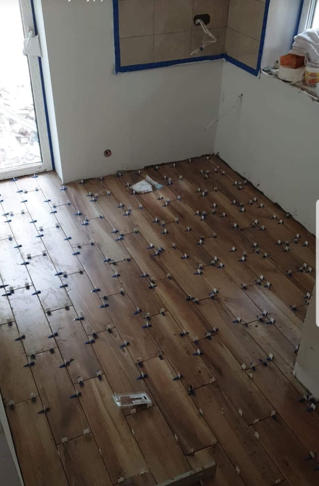 Układanie podłogi z drewnopodobnych paneli w trakcie prac wykończeniowych, widoczne krzyżyki dystansowe, ściany przygotowane do malowania, częściowo położone płytki na ścianie.