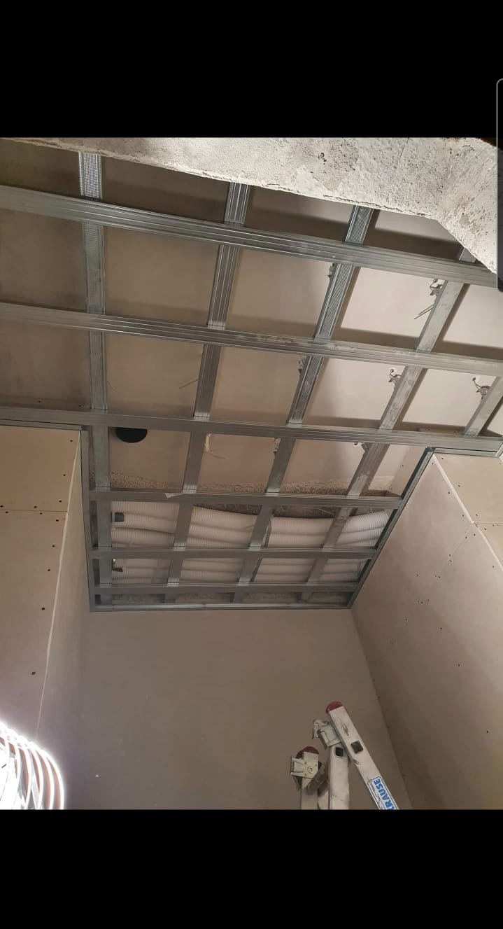 Montaż metalowej konstrukcji sufitu podwieszanego z widocznymi przewodami wentylacyjnymi, częściowo obłożonego płytami, widok z dołu z drabiny.