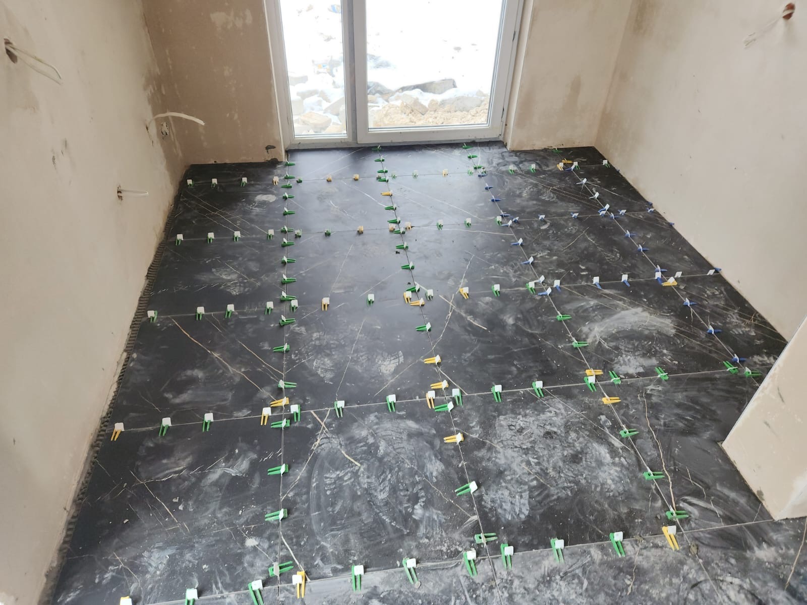 Układanie ciemnych płytek podłogowych imitujących marmur, z użyciem zielonych, żółtych i niebieskich klipsów poziomujących, widoczne surowe ściany i okno.