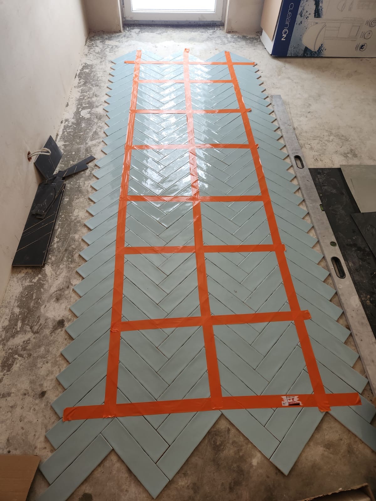 Układanie jasnoniebieskich płytek w jodełkę na podłodze, zabezpieczonych pomarańczową taśmą malarską, widoczne płytki do docinania przy ścianie i poziomnica.