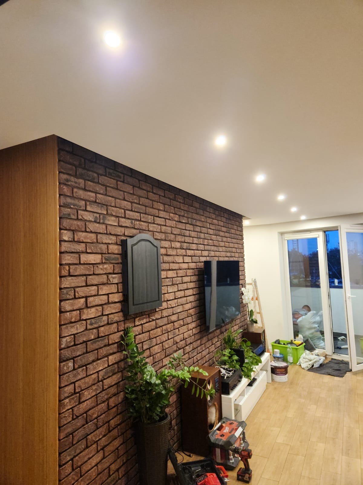 Salon z akcentem ceglanej ściany, drewnianą szafką po lewej, roślinami doniczkowymi, sprzętem audio i narzędziami, widoczne przesuwne drzwi balkonowe i osoba w tle.