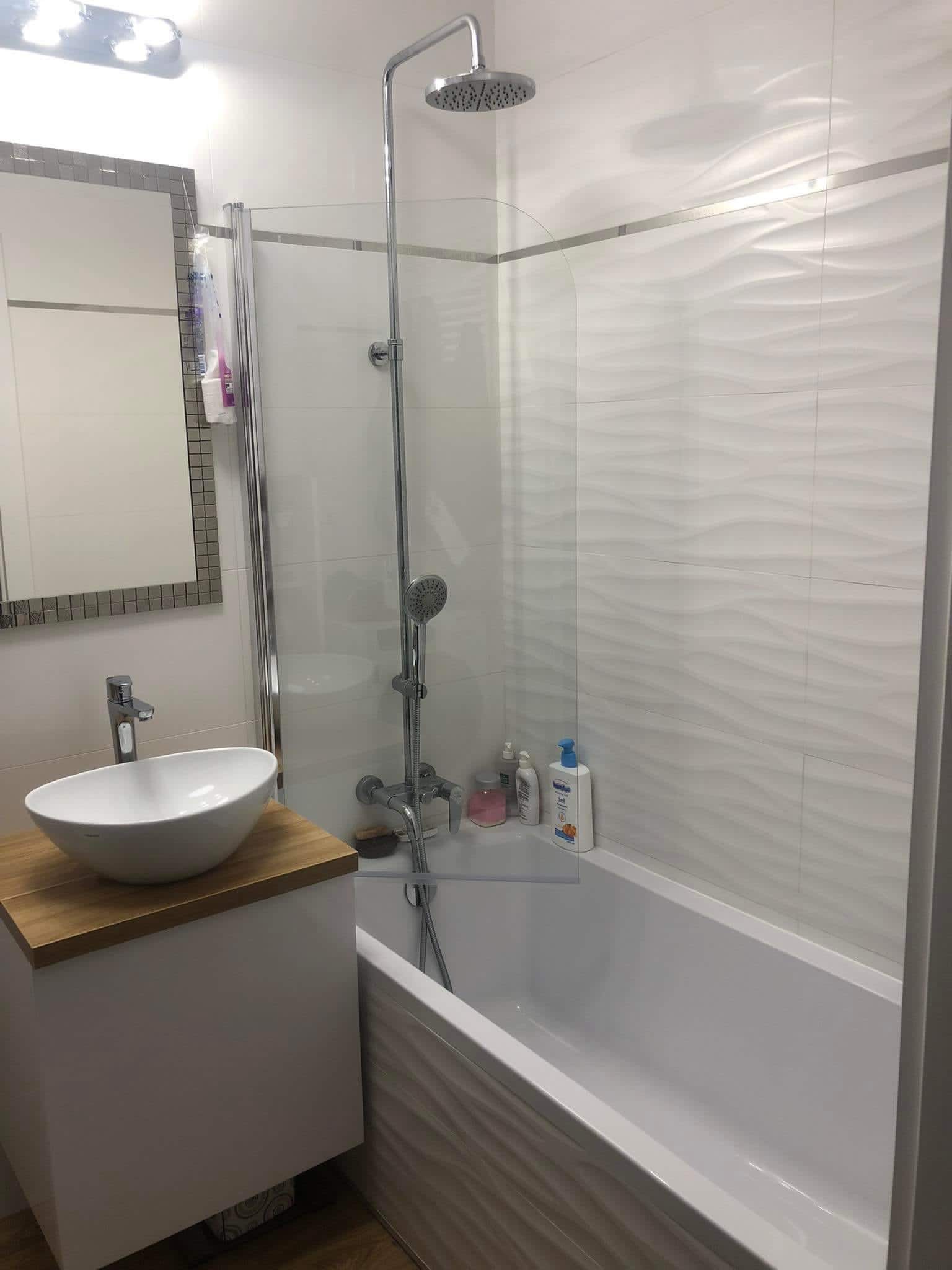 Nowoczesna łazienka z białą armaturą, umywalką nablatową na drewnianym blacie, kabiną prysznicową z hydromasażem i płytkami z delikatnym wzorem fali. Oświetlenie sufitowe i lustro z mozaikową ramą...