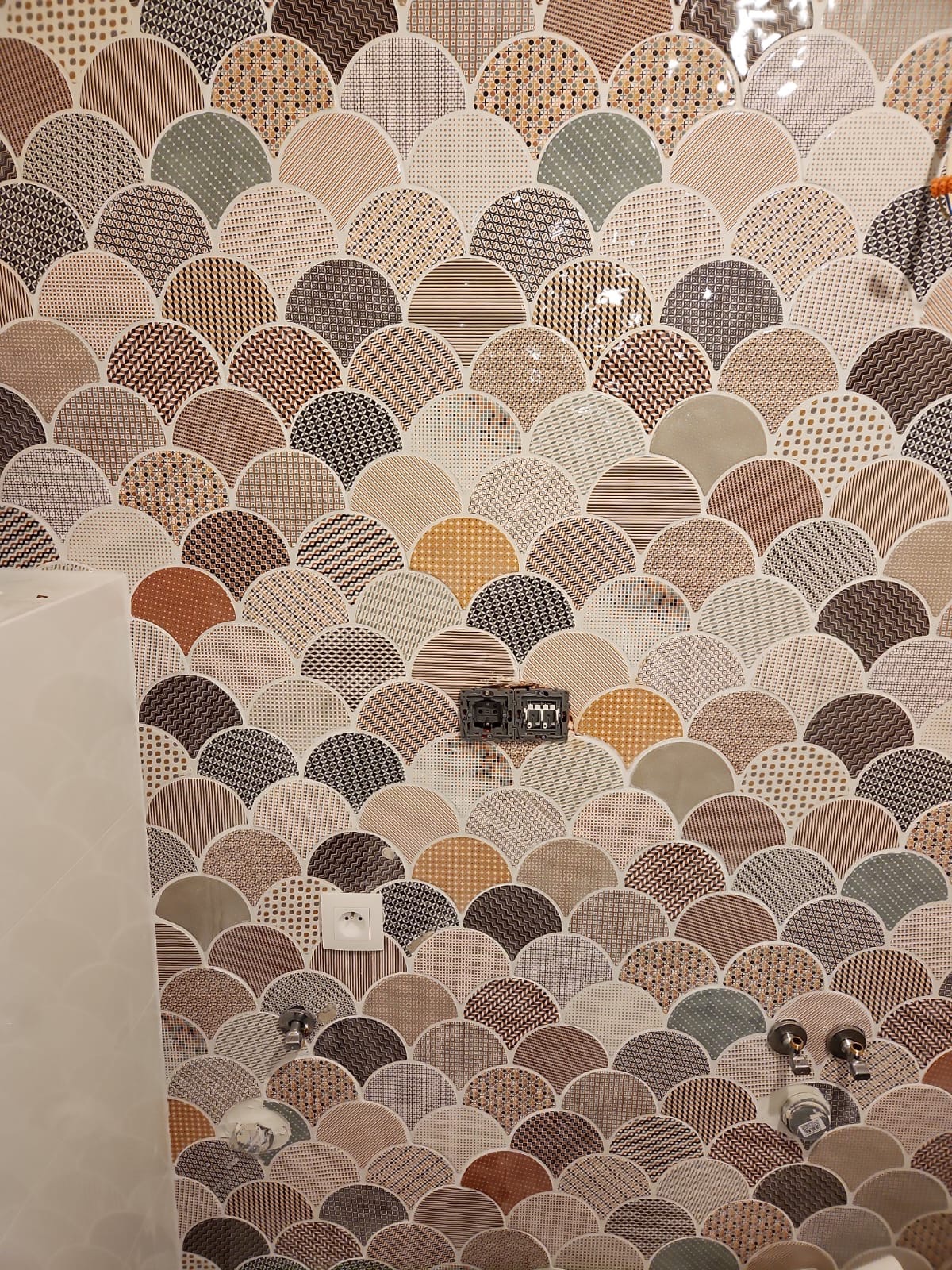 Ściana wyłożona dekoracyjnymi płytkami ceramicznymi w kształcie rybiej łuski w odcieniach brązu i beżu, z widocznymi gniazdkami elektrycznymi i przyłączami wodnymi, sugerująca etap prac podczas...