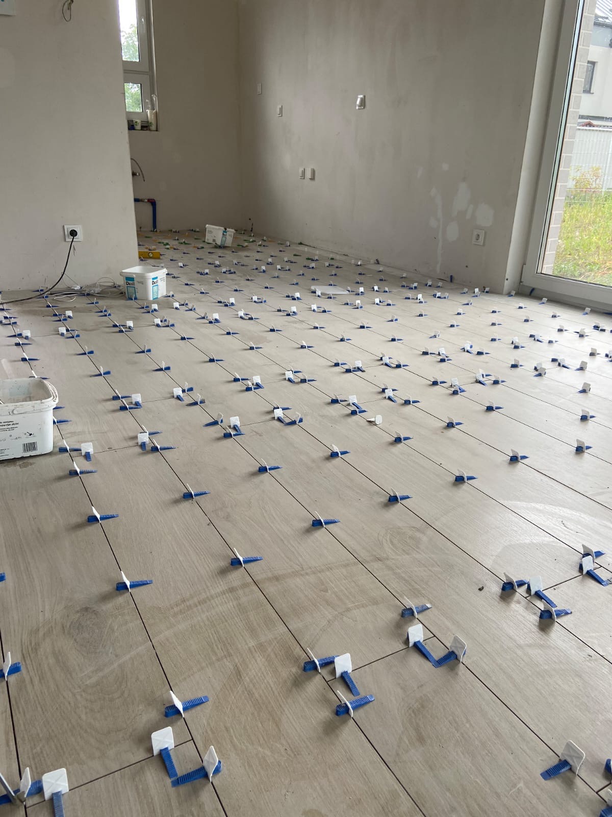 Układanie płytek podłogowych w jasnym odcieniu drewna, z użyciem niebieskich krzyżyków dystansowych, widoczne wiaderka z klejem i surowe ściany w tle.