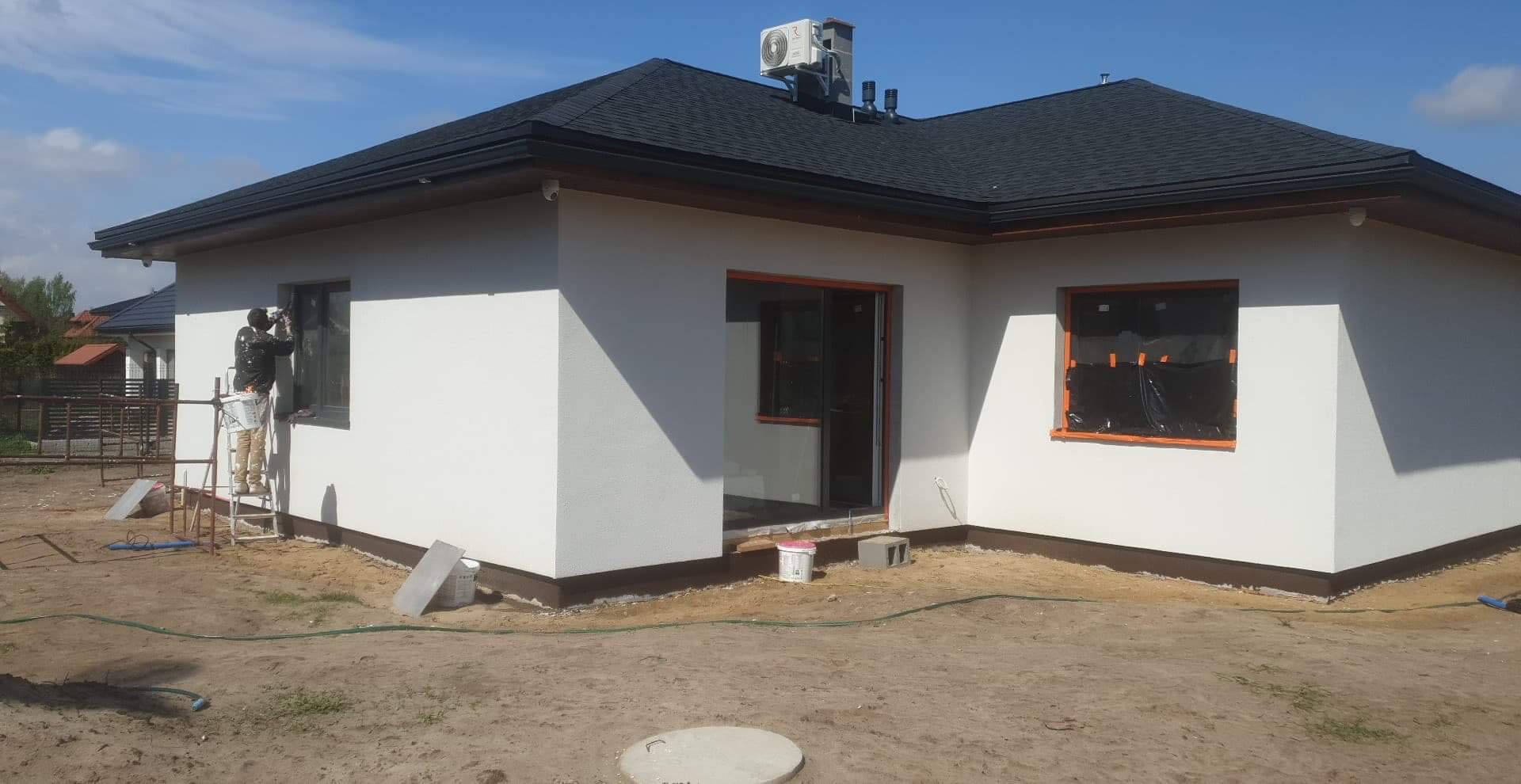 Malowanie elewacji domu jednorodzinnego z czarnym dachem, widoczny pracownik na drabinie malujący ramę okienną na szaro, okno zabezpieczone folią, obok stoi wiadro z farbą.