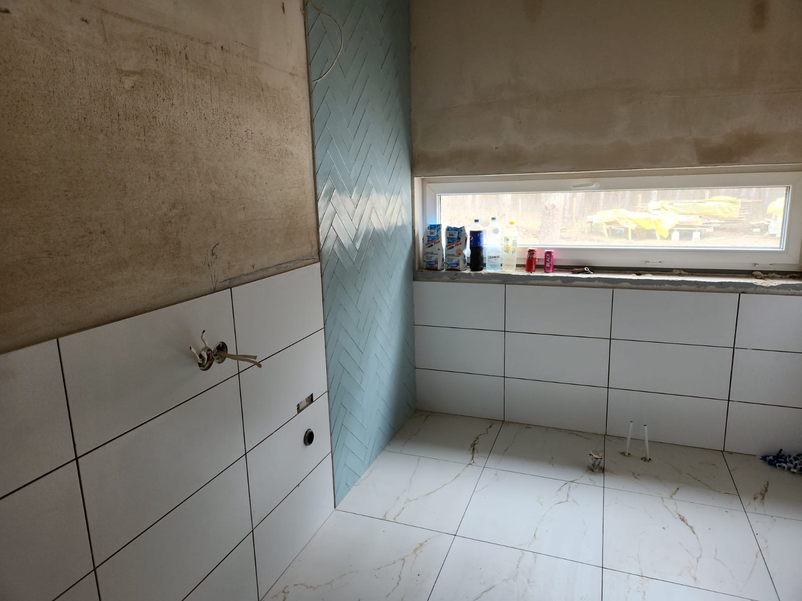 Remontowana łazienka w Warszawie: ściany częściowo wyłożone białymi płytkami, fragment ściany z niebieską mozaiką ułożoną w jodełkę, surowy fragment ściany z widocznym tynkiem, marmurowe płytki...