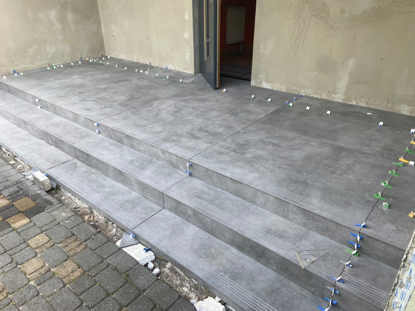 Układanie szarych płytek na schodach wejściowych, widoczne plastikowe krzyżyki dystansowe i fragment kostki brukowej.