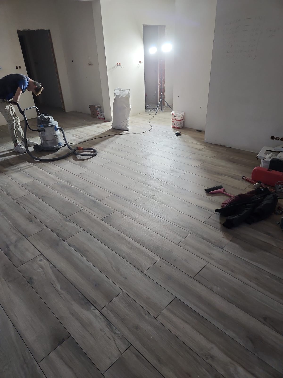 Wnętrze w trakcie remontu: mężczyzna odkurza nowo ułożoną podłogę z płytek imitujących drewno, widoczne worki z materiałami budowlanymi, oświetlenie robocze i ślady po pracach na ścianach.