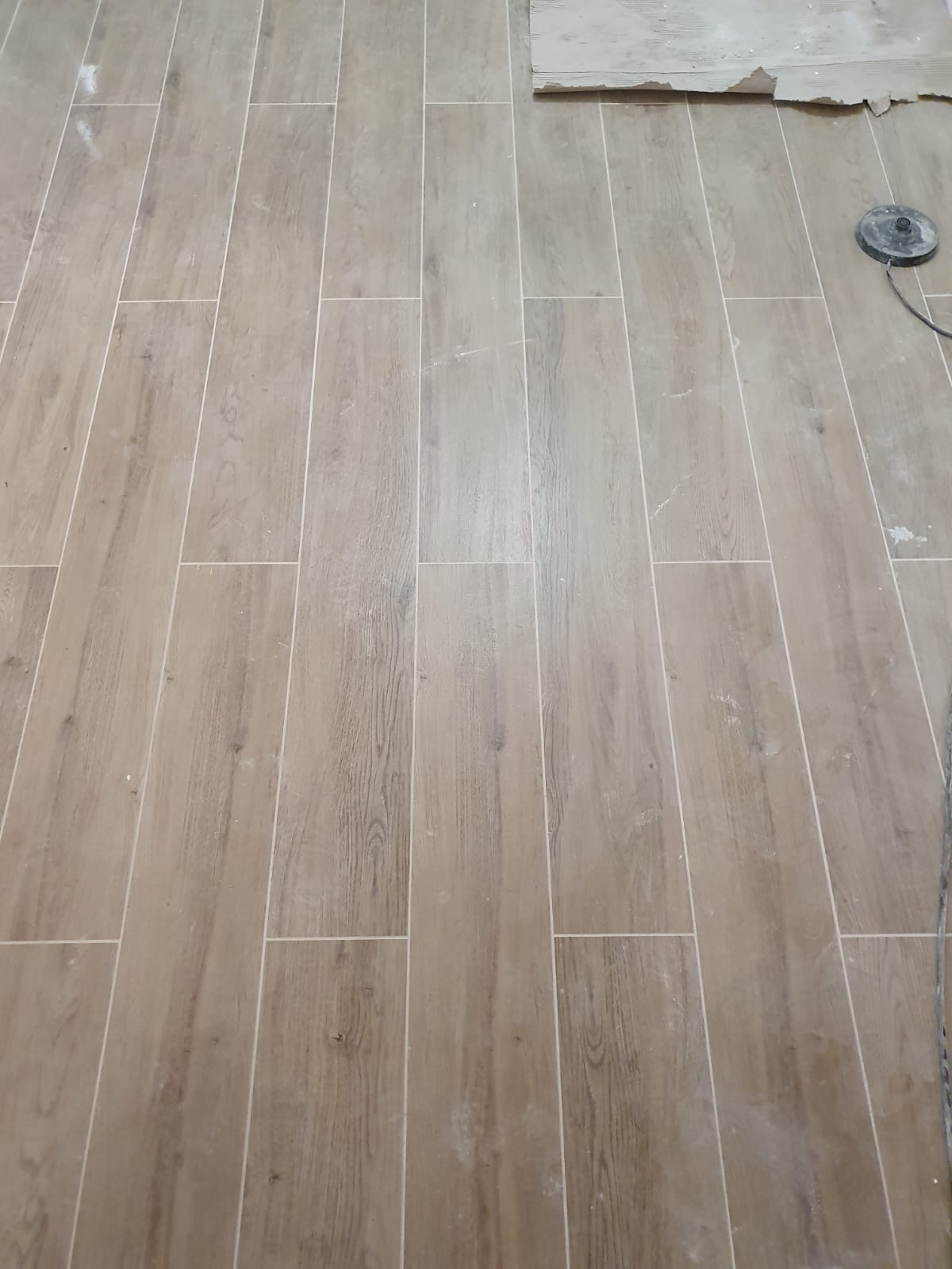Ułożone podłużne panele podłogowe imitujące drewno, widoczne białe fugi, pozostałości zaprawy i narzędzie szlifierskie w rogu.