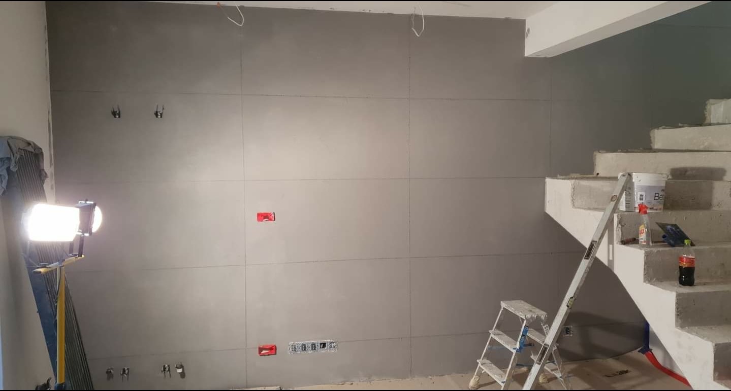 Ściana z płyt gipsowo-kartonowych w trakcie prac wykończeniowych, widoczne puszki elektryczne, przewody, drabina, poziomica i akcesoria malarskie na schodach w surowym stanie.