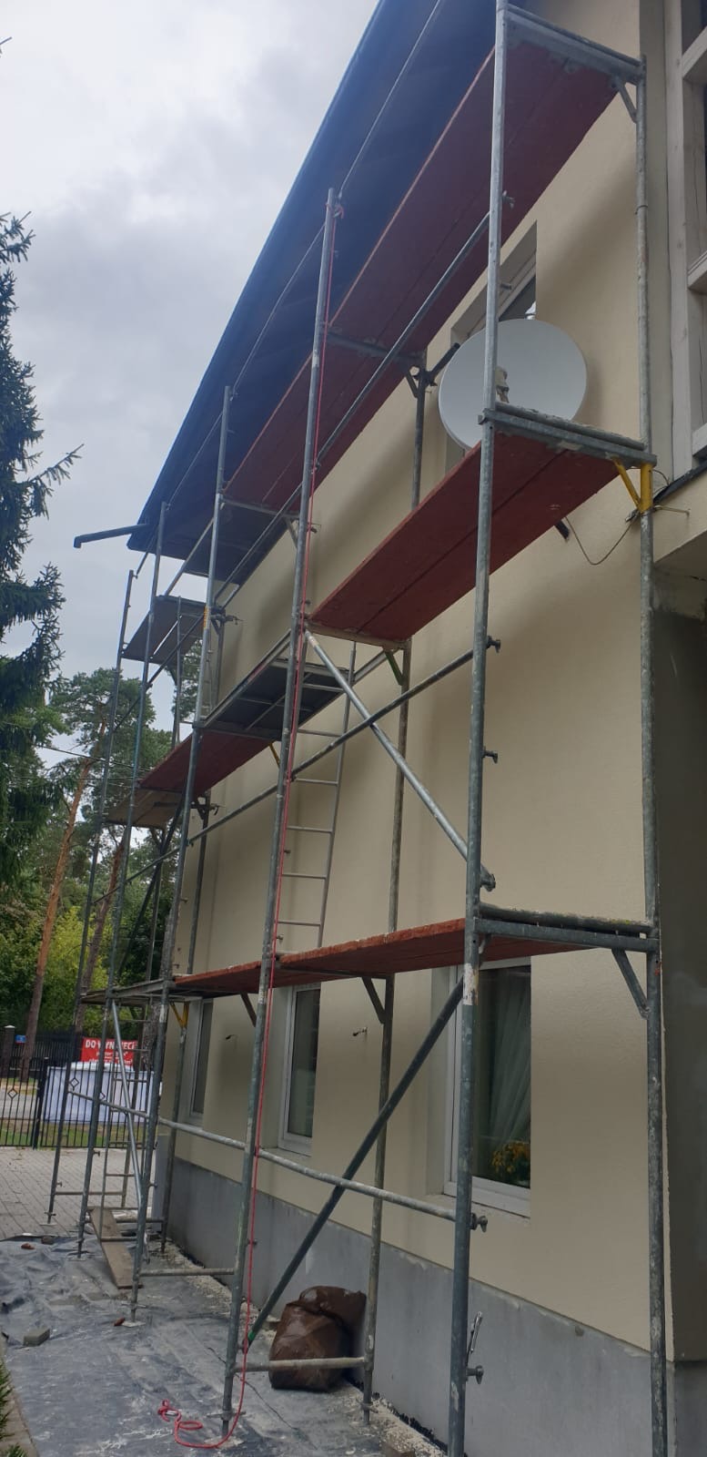 Elewacja budynku w trakcie malowania, widoczne rusztowania, pomalowana górna część ściany na beżowo, dolna część w szarym podkładzie, antena satelitarna na ścianie.