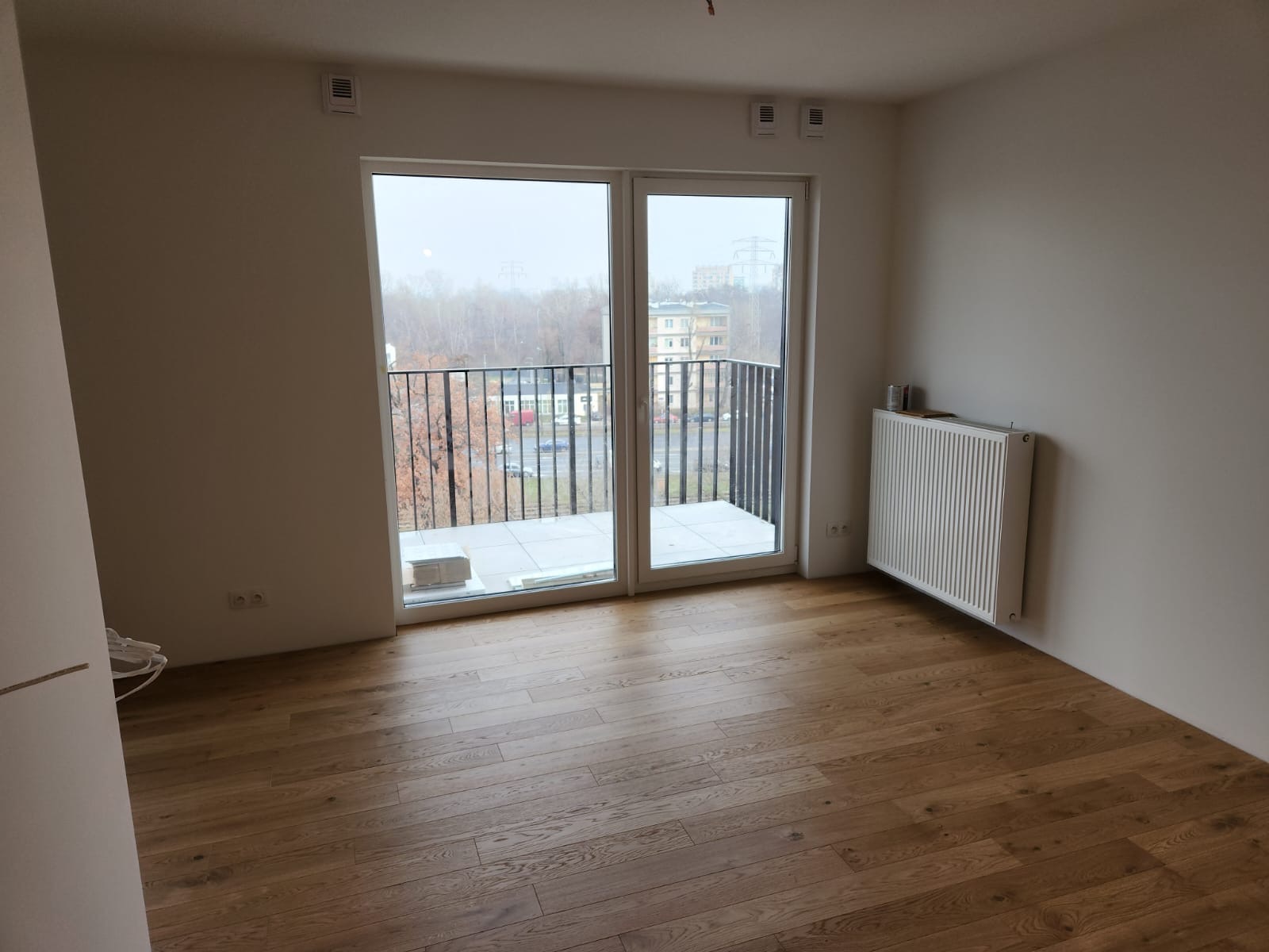 Pusty pokój z drewnianą podłogą, białymi ścianami i balkonem z metalową balustradą. Na balkonie widoczne białe płytki i pakiet materiałów budowlanych. Grzejnik zamontowany na ścianie. Widok...