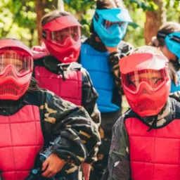 Grupa dzieci w maskach i kamizelkach ochronnych, ubranych w odzież moro, przygotowana do gry w paintball w lesie.