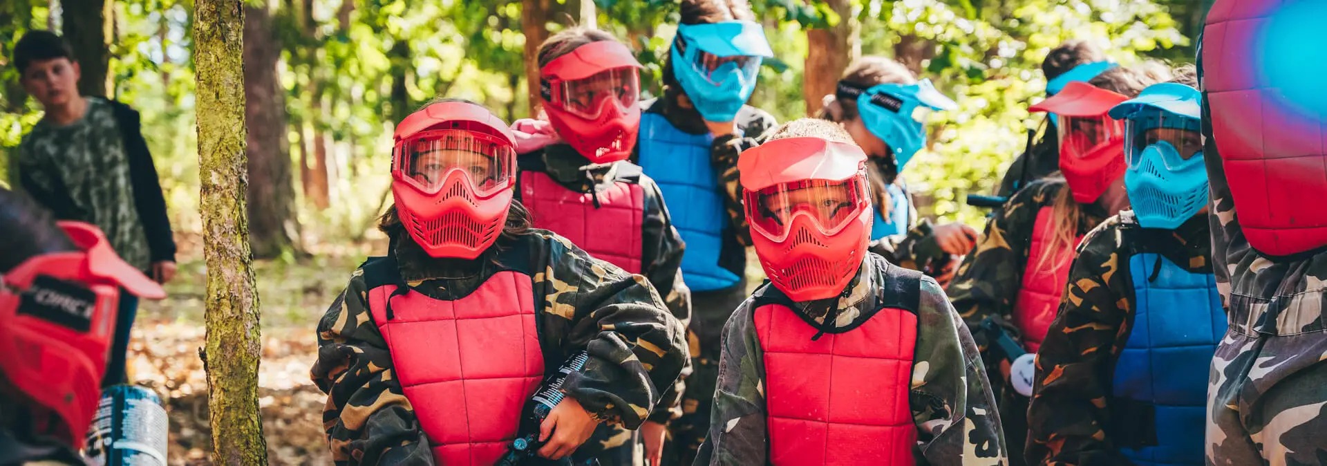 Grupa dzieci w maskach i kamizelkach ochronnych, ubranych w odzież moro, przygotowana do gry w paintball w lesie.