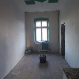 Pokój w trakcie remontu: świeżo otynkowane ściany, odsłonięta futryna drzwiowa, stare drewniane podłogi z resztkami farby, okno zabezpieczone folią, dekoracyjny kaseton na suficie.