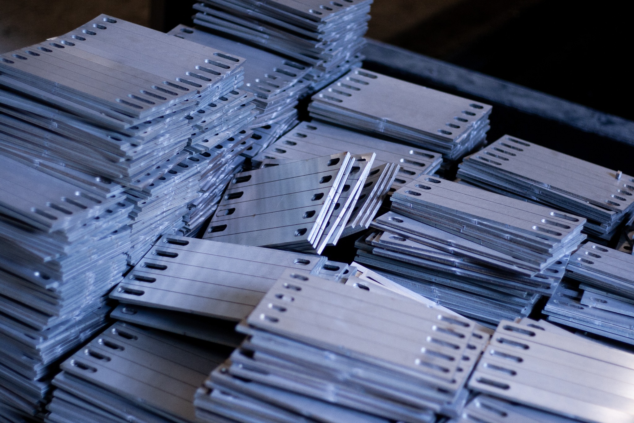 Stosy srebrnych, metalowych płyt montażowych z otworami i szczelinami, prawdopodobnie do konstrukcji wsporczych paneli słonecznych, ułożone w nieładzie.