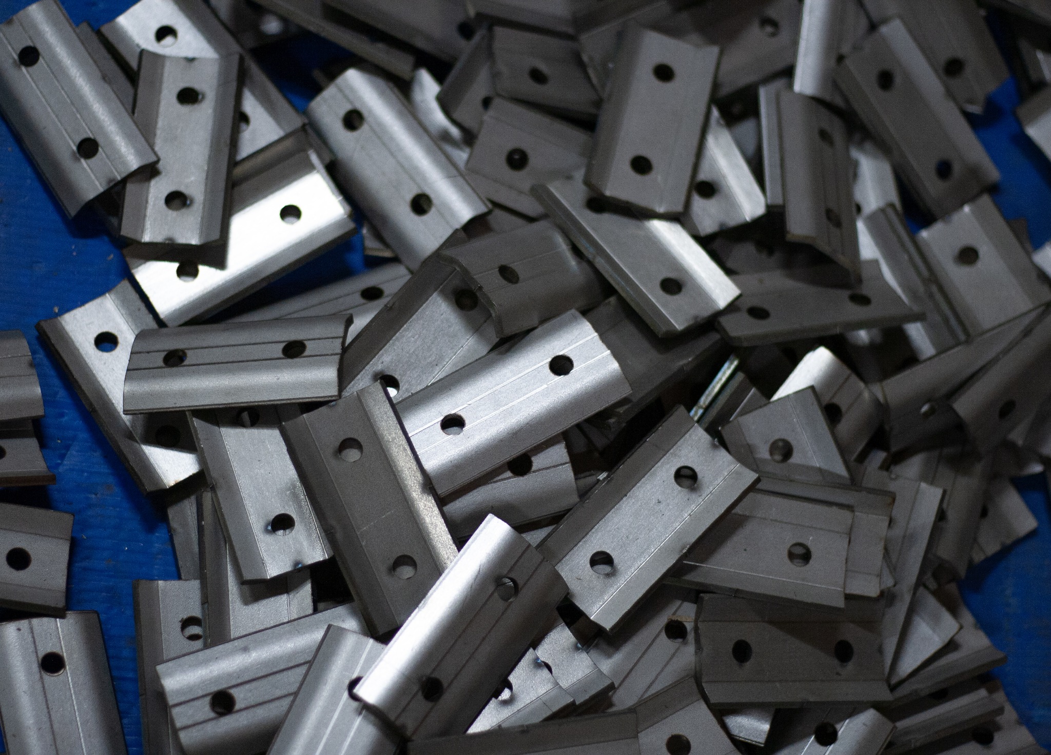 Stalowe uchwyty montażowe z dwoma otworami ułożone chaotycznie na niebieskim tle.