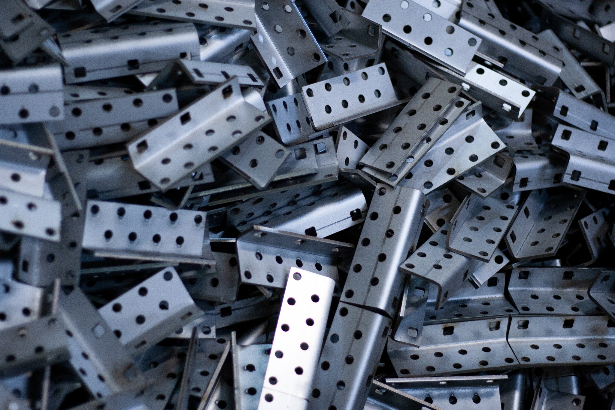 Stalowe, perforowane kątowniki montażowe do konstrukcji, ułożone chaotycznie w dużym skupisku, z widocznymi otworami o różnych średnicach i kształtach.