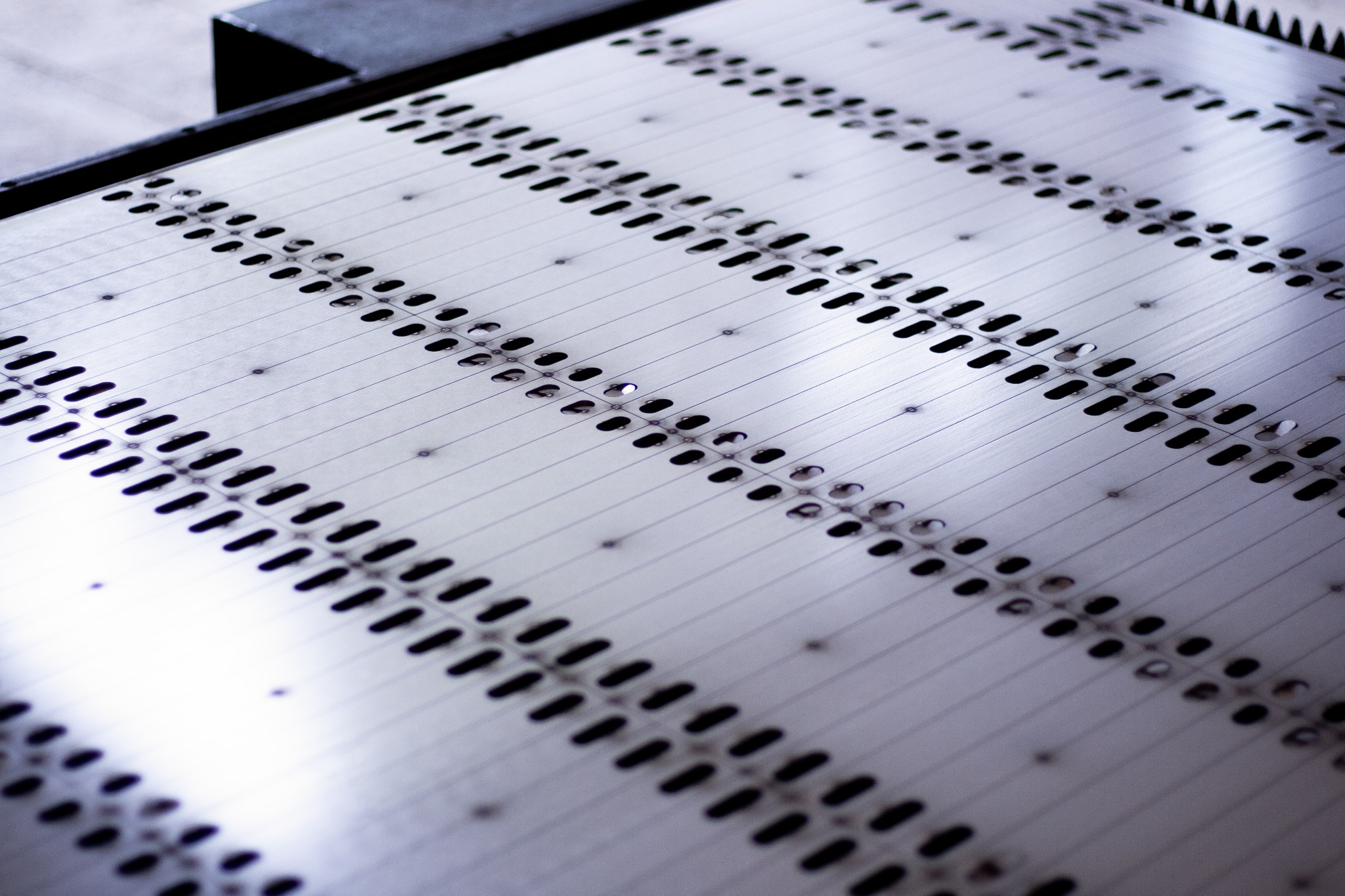 Metalowa płyta z rzędami otworów, prawdopodobnie element konstrukcyjny modułu fotowoltaicznego, widoczne drobne nity i refleksy światła.