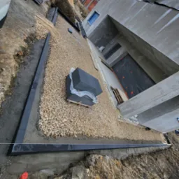 Przygotowanie podłoża pod taras: wysypany żwir, ułożone obrzeża z kostki betonowej, widoczny budynek w trakcie budowy w tle.