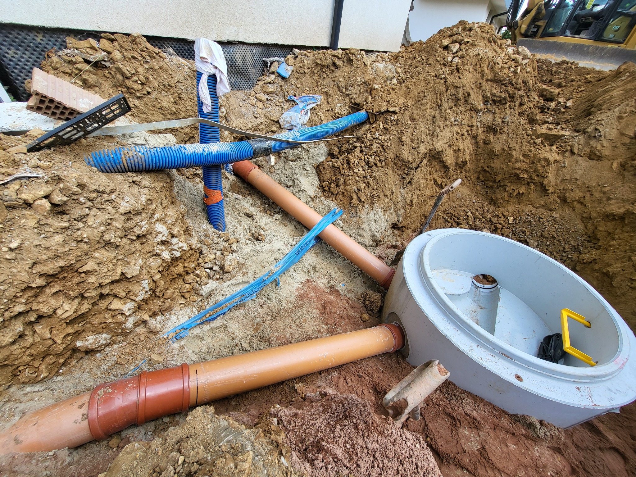 Wykonany wykop z widocznymi rurami kanalizacyjnymi z tworzywa sztucznego, studzienką rewizyjną z żółtym uchwytem w środku i ziemią, w tle ściana budynku i koparka.