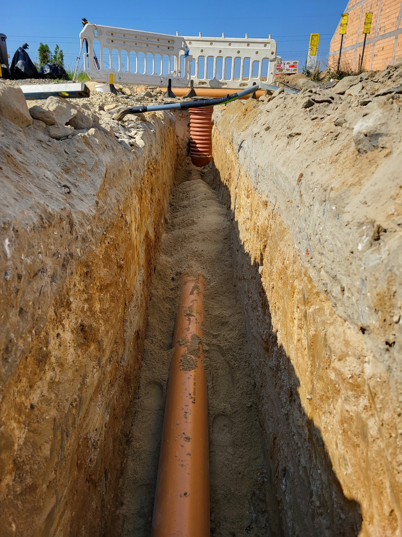 Wykop w ziemi z ułożonymi rurami kanalizacyjnymi, widoczny fragment zabezpieczenia wykopu i oznakowanie robót.