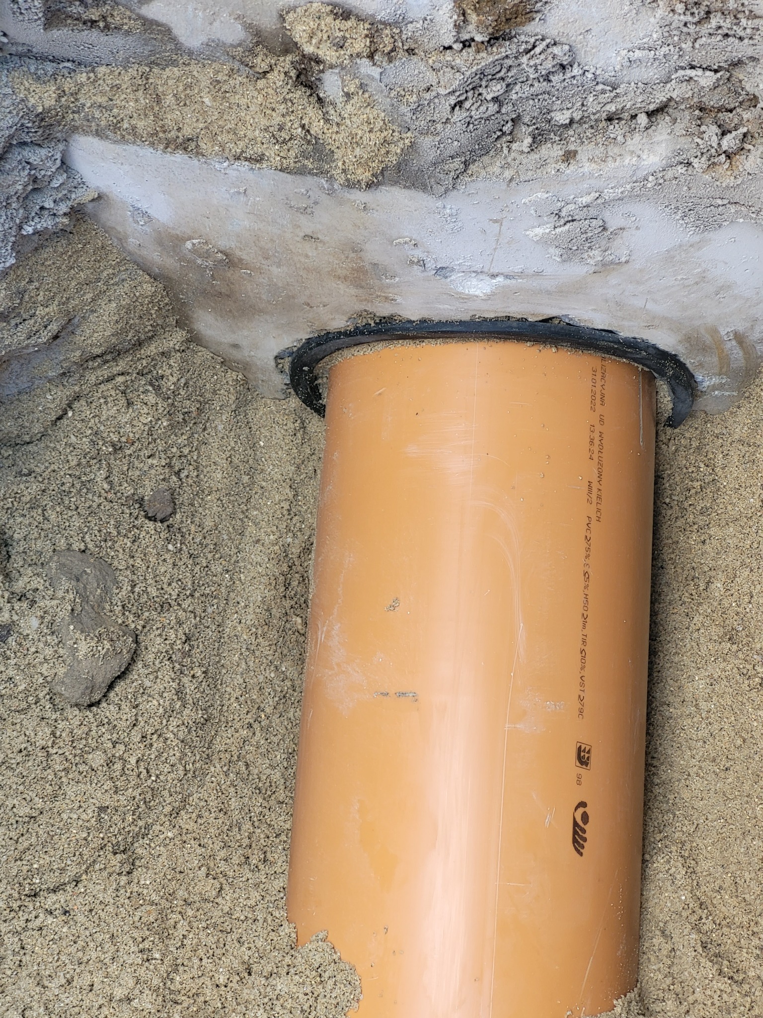 Pomarańczowa rura kanalizacyjna wystająca z gruntu, uszczelniona czarną gumową uszczelką, otoczona piaskiem i betonem.