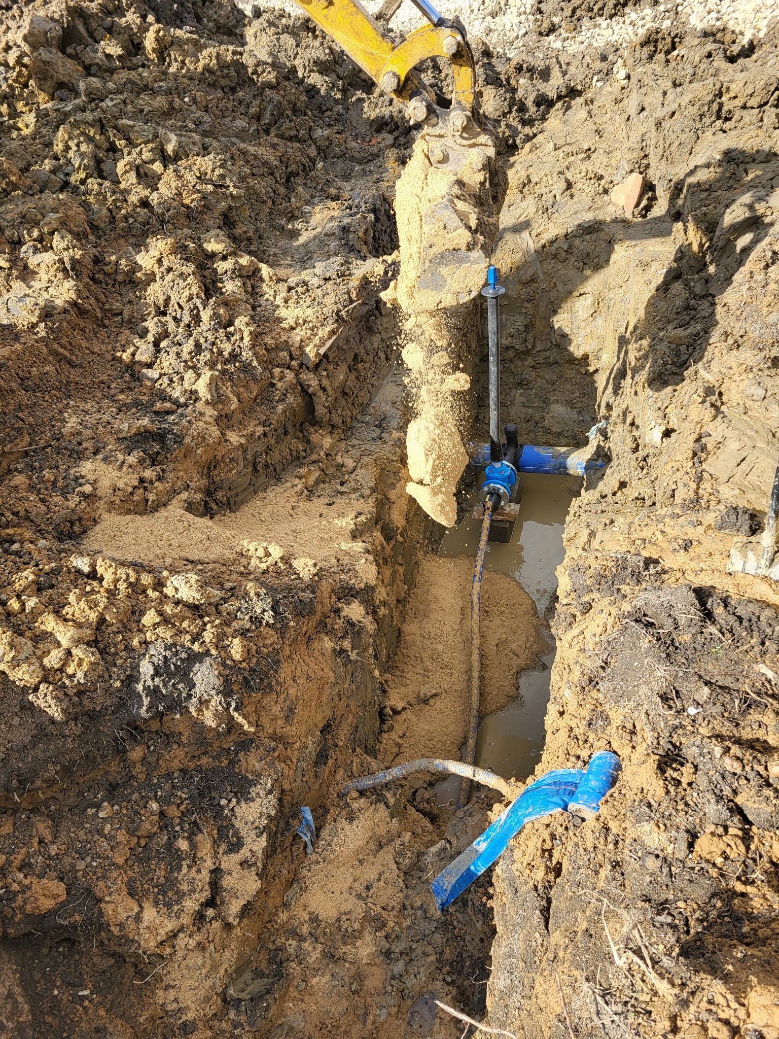 Wykop pod przyłącze wodno-kanalizacyjne z widocznymi rurami, złączkami i żółtą łyżką koparki w tle, częściowo wypełniony wodą i piaskiem.