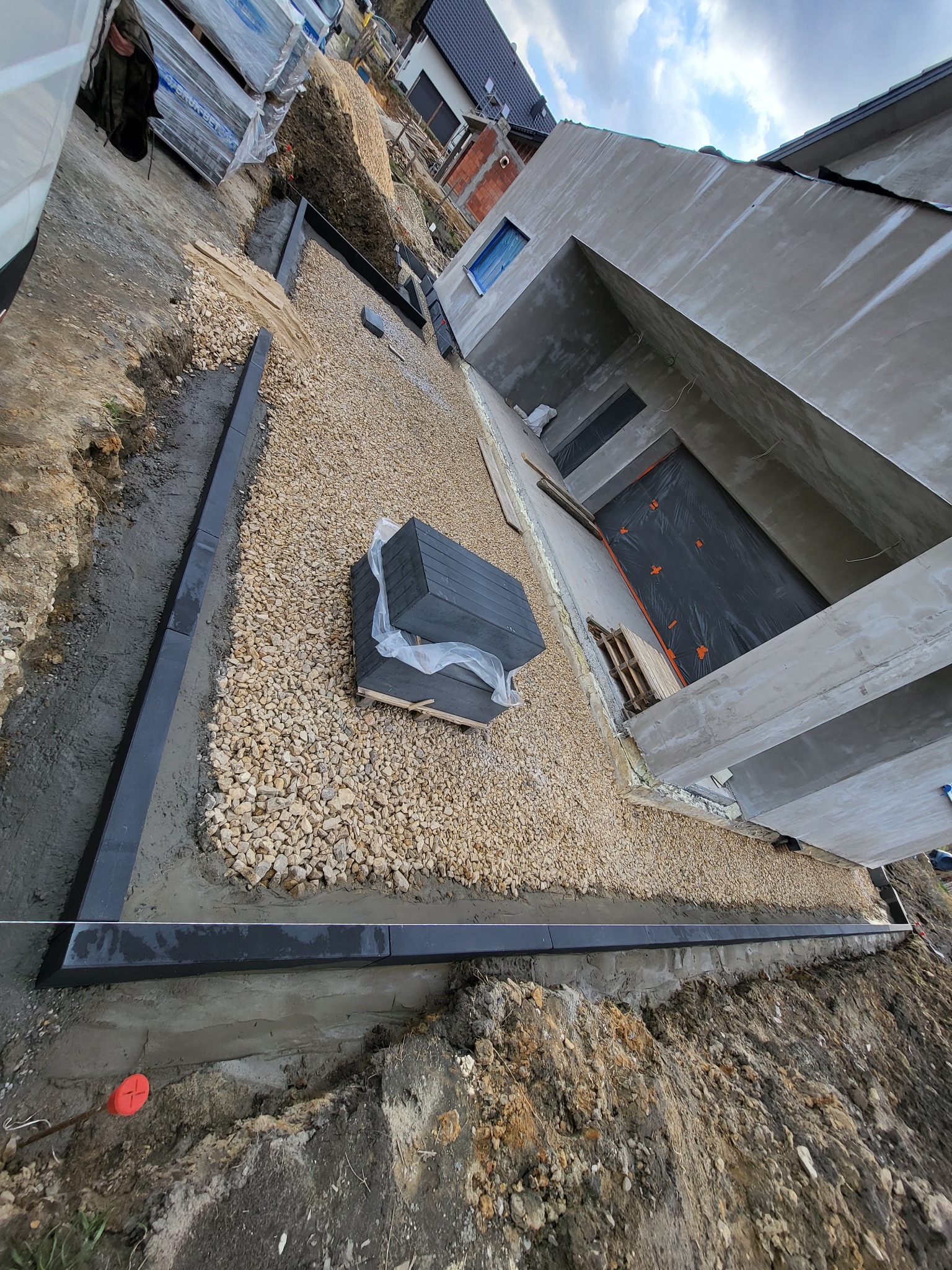 Przygotowanie podłoża pod taras: wysypany żwir, ułożone obrzeża z kostki betonowej, widoczny budynek w trakcie budowy w tle.