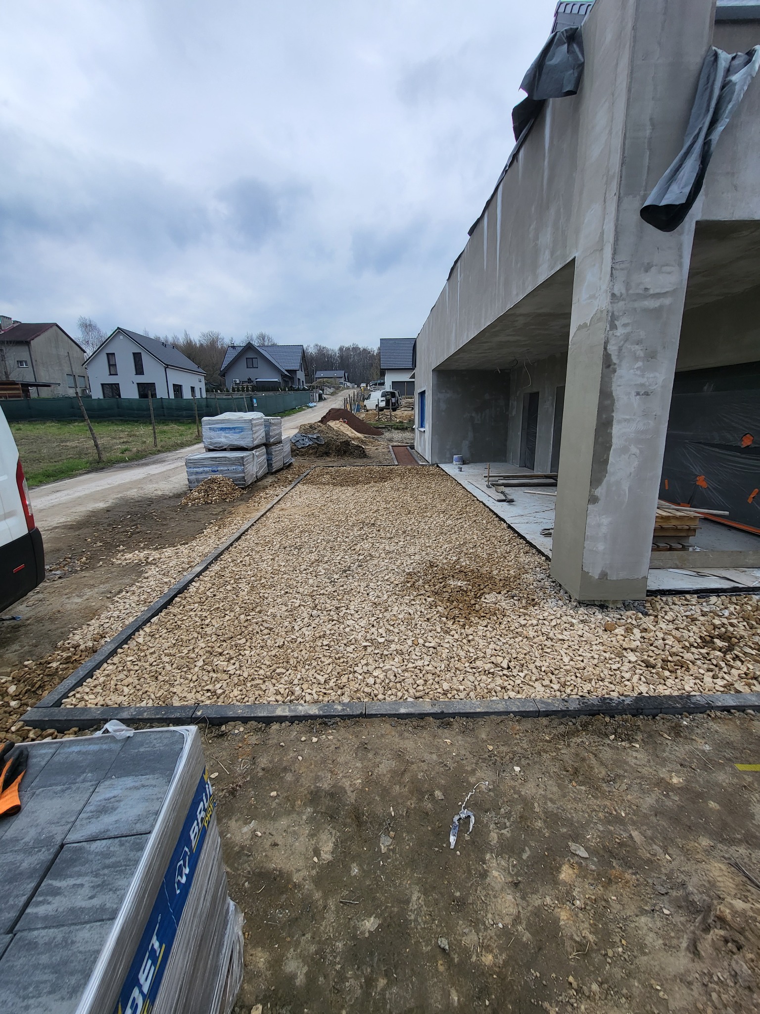 Przygotowany teren pod budowę podjazdu z wysypanym żwirem, obrzeża ułożone, w tle budynek w trakcie wykańczania elewacji i sterta kostki brukowej w folii.