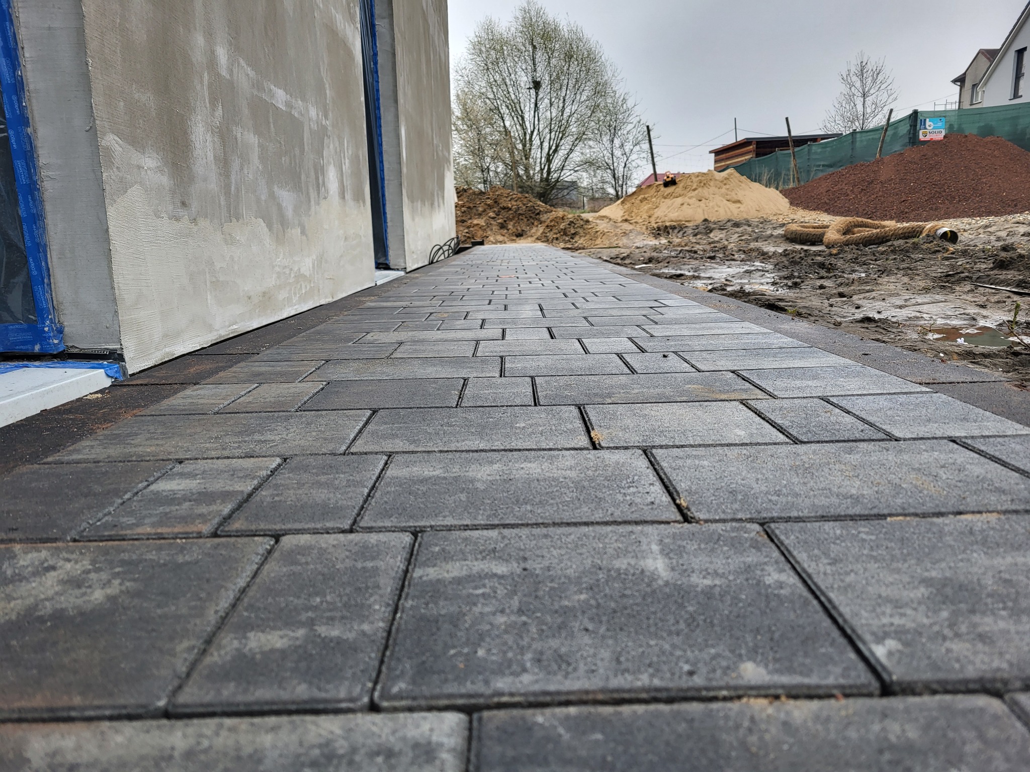 Świeżo ułożona, szara kostka brukowa prowadząca wzdłuż ściany budynku, w tle hałdy piasku i ziemi oraz drzewo bez liści pod pochmurnym niebem.