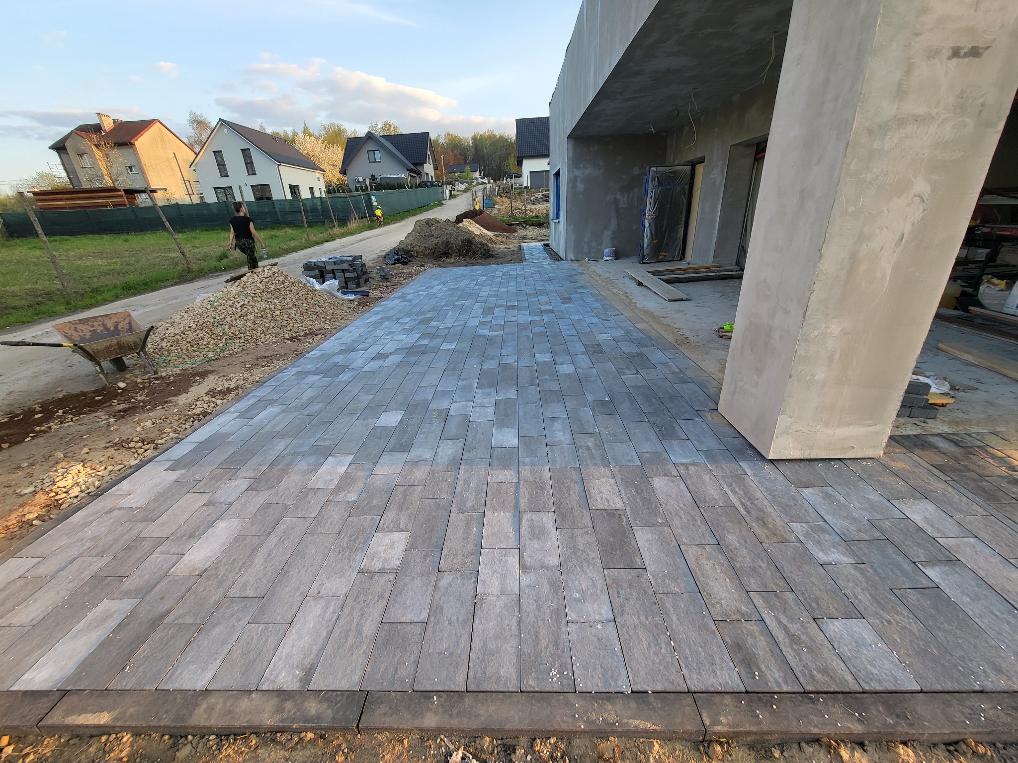 Nowo ułożona, szara kostka brukowa prowadząca do surowego budynku w trakcie budowy, widoczny stos kostki i taczka z materiałem budowlanym w tle.