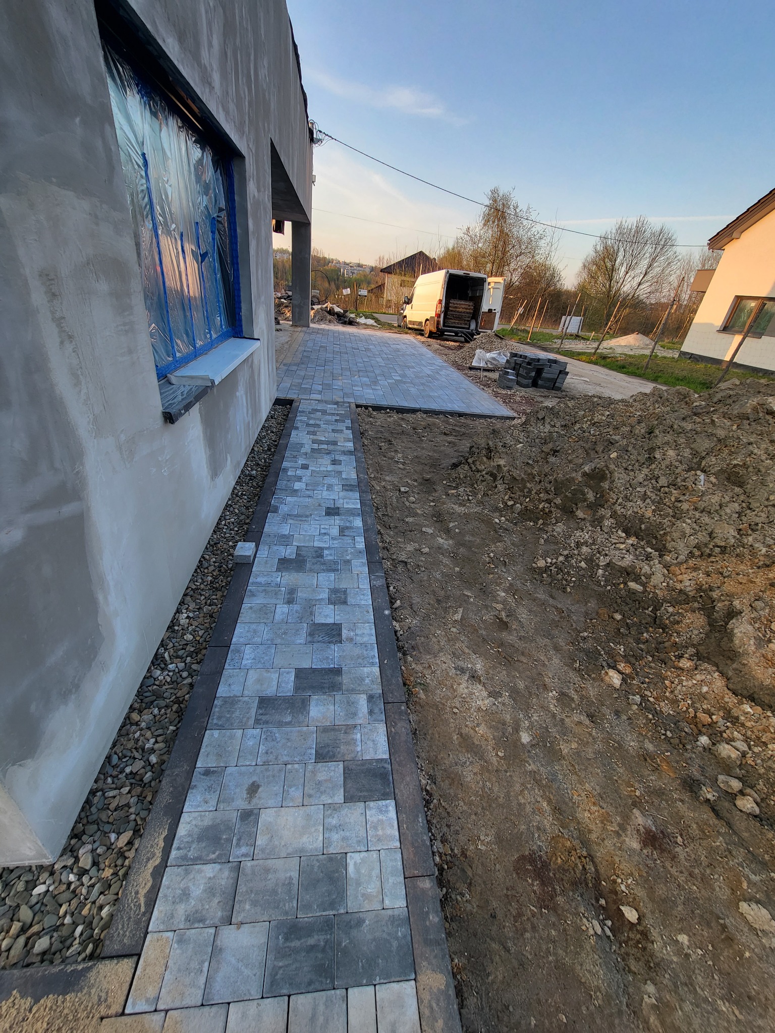 Ścieżka z szarej kostki ułożona wzdłuż surowej ściany budynku, obok wąski pas z kamykami, w tle zaparkowany biały bus i sterta ziemi.