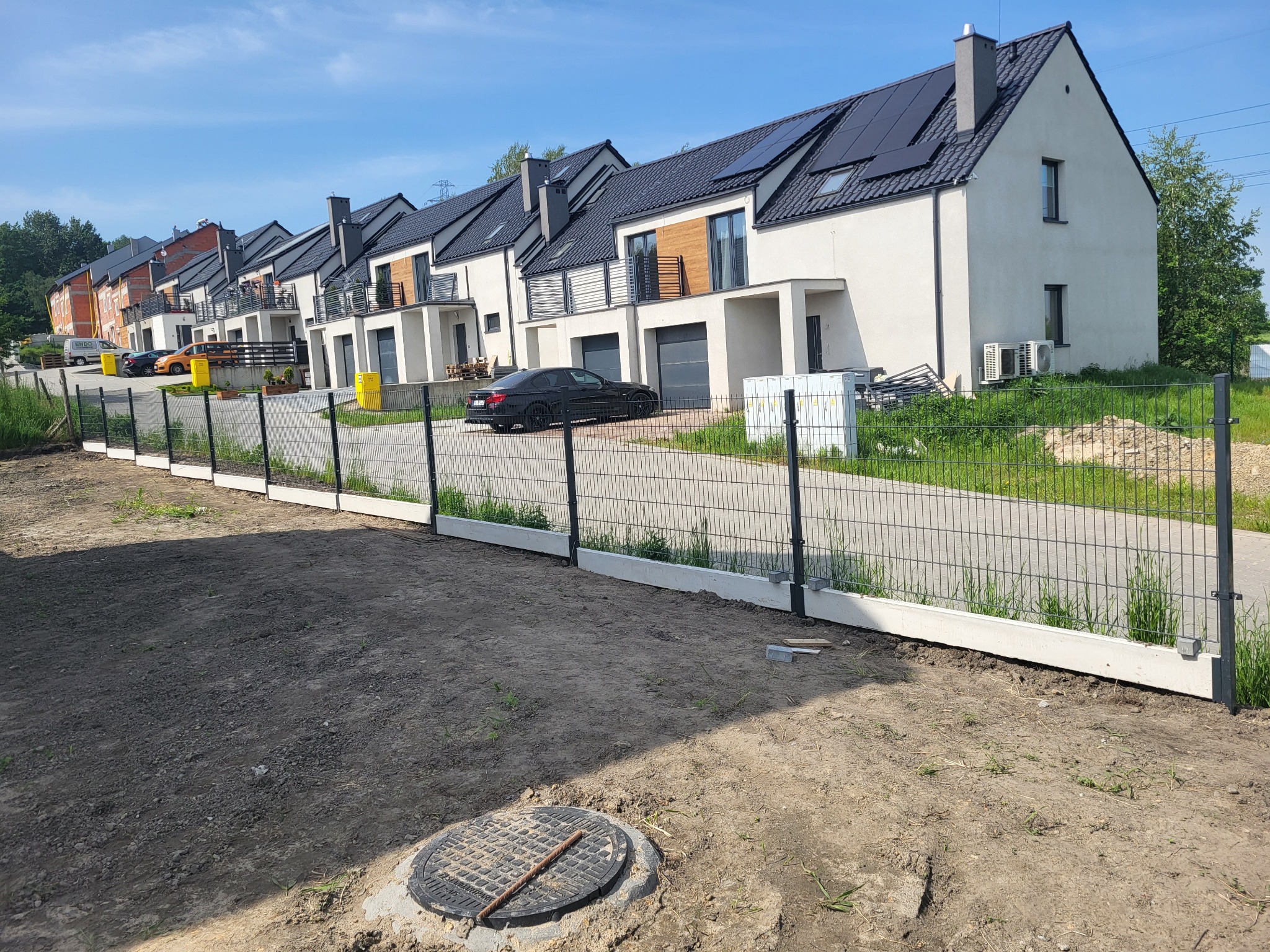 Nowoczesne szeregowe domy z panelami słonecznymi, widoczne zza metalowego ogrodzenia z betonową podmurówką na jeszcze niezagospodarowanym terenie.