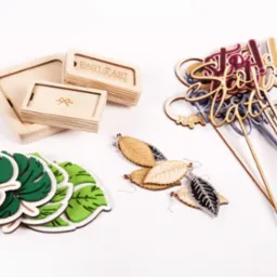 Ręcznie robione, drewniane i filcowe ozdoby: pudełka z logo, kolczyki w kształcie liści, dekoracje na piku z napisami na białym tle.