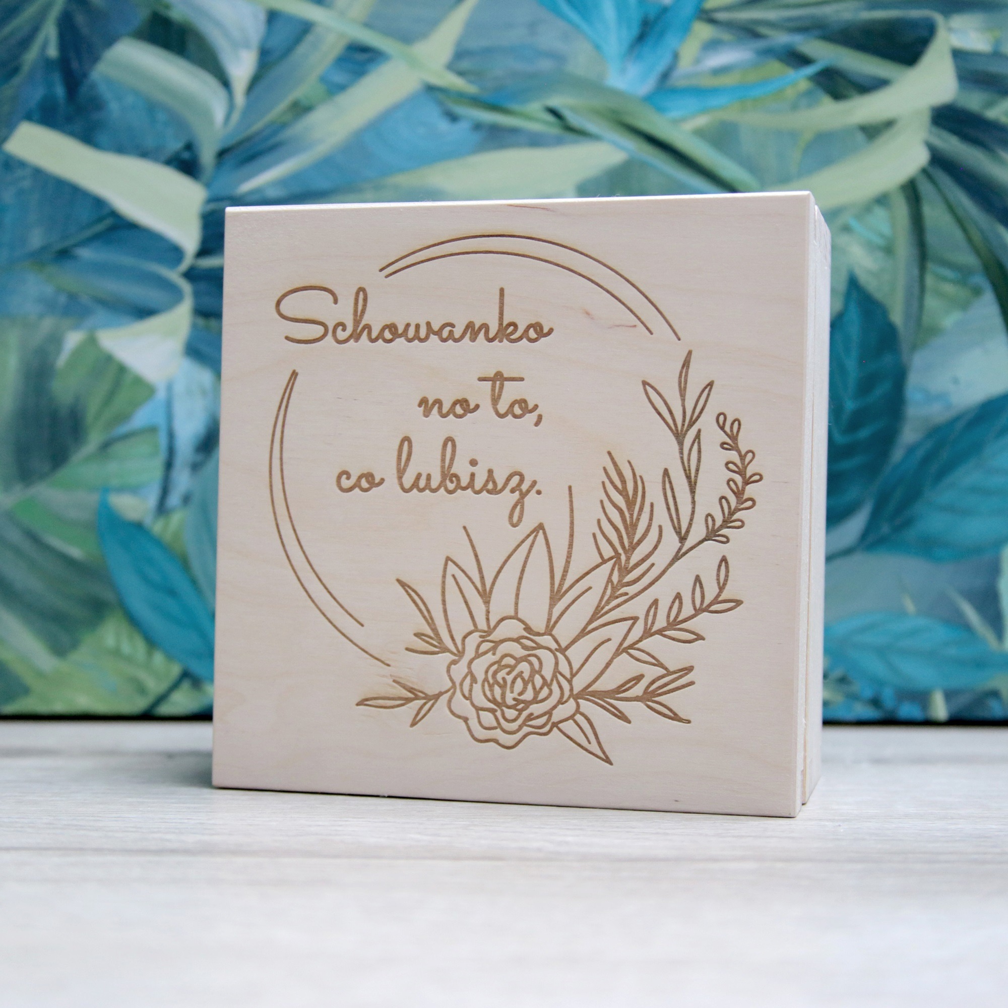 Drewniane pudełko z wygrawerowanym napisem 'Schowanko na to, co lubisz' i motywem kwiatowym na tle abstrakcyjnego obrazu w odcieniach zieleni i błękitu.
