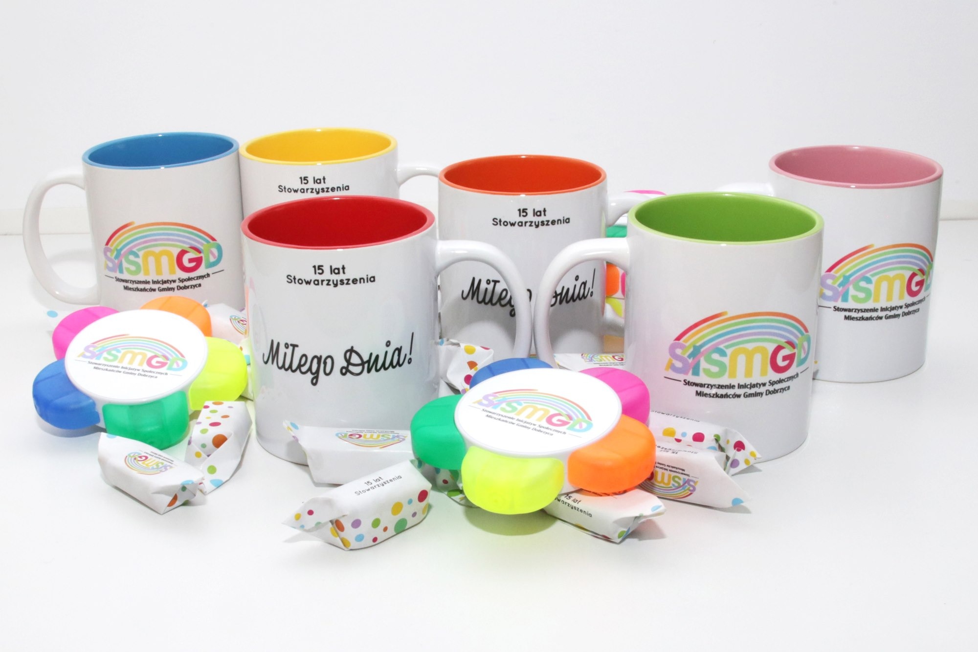 Zestaw upominkowy z białych kubków z kolorowym wnętrzem i nadrukiem logo oraz napisem '15 lat Stowarzyszenia' i 'Miłego Dnia!', uzupełniony o kolorowe gadżety i słodycze w ozdobnych papierkach.