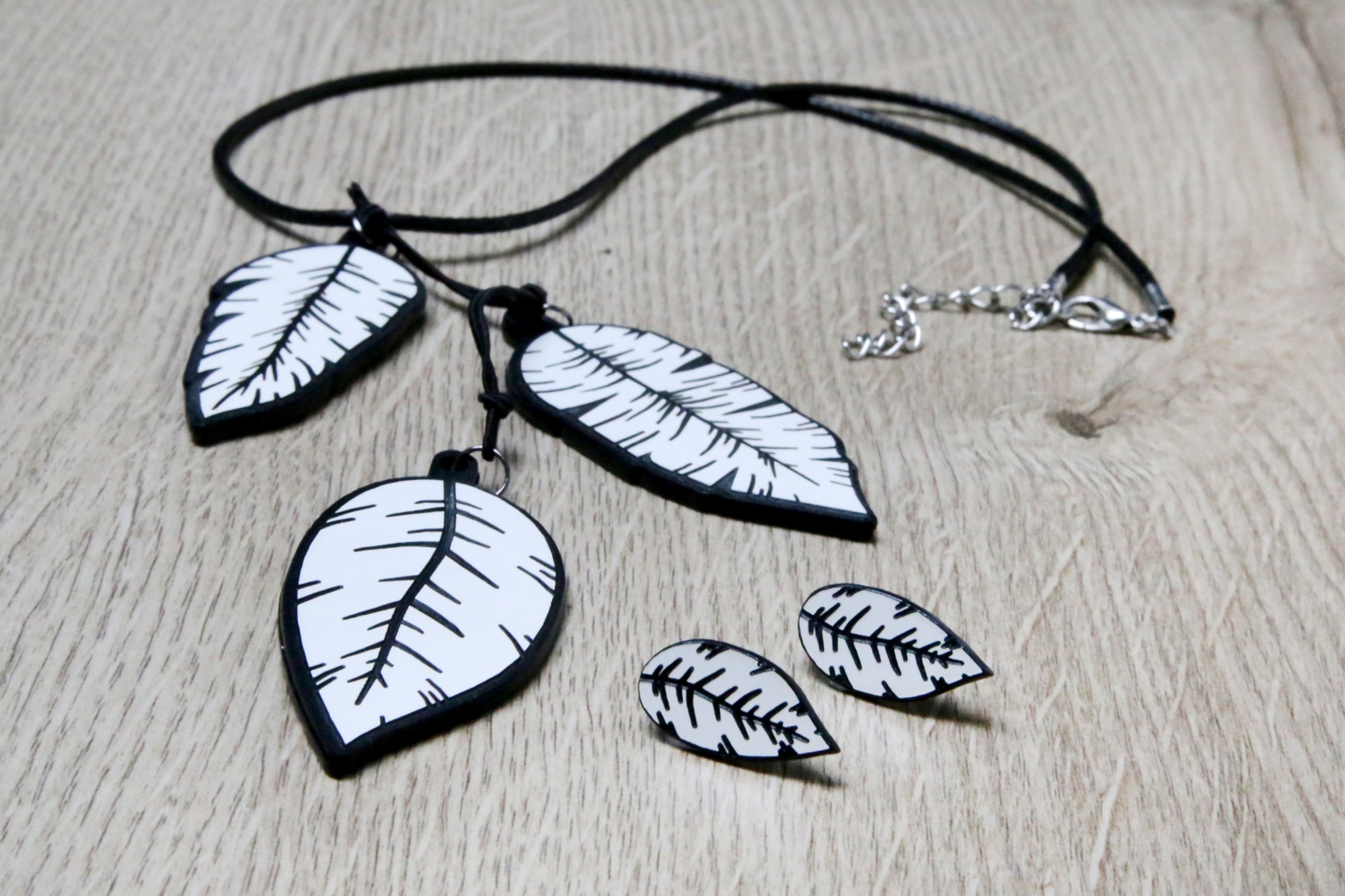 Ręcznie robiony komplet biżuterii: naszyjnik z trzema zawieszkami w kształcie liści i para kolczyków, czarno-białe, na drewnianym tle.