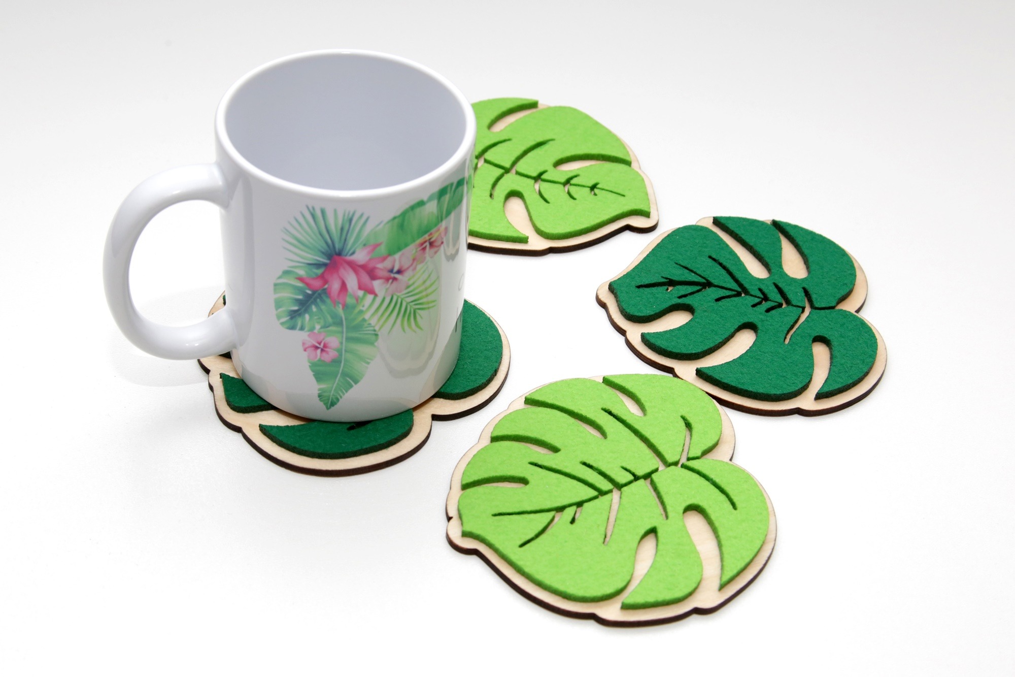 Biały kubek z motywem tropikalnym na podkładce w kształcie liścia monstery, obok trzy identyczne podkładki filcowo-drewniane na białym tle.