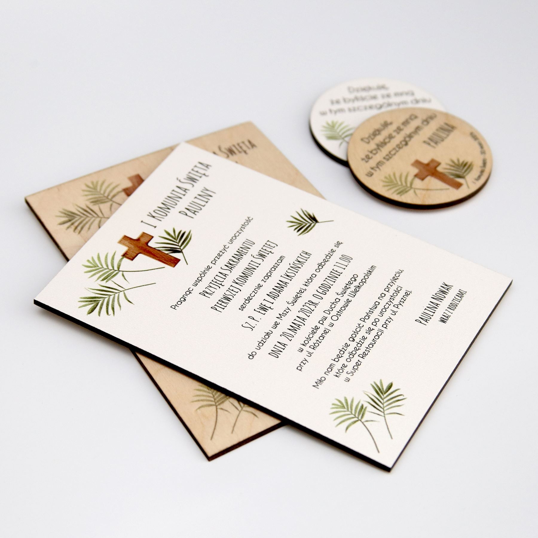 Zaproszenie na Pierwszą Komunię Świętą Pauliny z drewnianymi elementami dekoracyjnymi, z motywem krzyża i liści palmowych, leżące na białym tle.