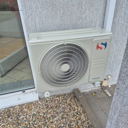 Arkadiusz Zaustowicz Klima Serwis - Zamontowana jednostka zewnętrzna klimatyzatora Sinclair na elewacji budynku, widoczne kamyki dekoracyjne i fragment okna z odbiciem wnętrza.