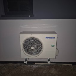 Arkadiusz Zaustowicz Klima Serwis - Zewnętrzna jednostka klimatyzacji Panasonic zamontowana na wspornikach przy szarej ścianie, widoczny fragment okna w ciemnej ramie i niedokończona podłoga.
