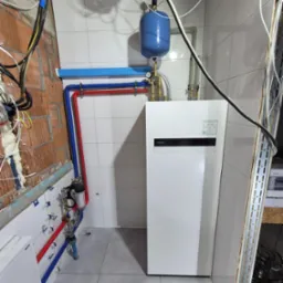 Instalacja pompy ciepła Panasonic Aquarea Smart Cloud w pomieszczeniu z częściowo wykończonymi ścianami, widoczne rury w kolorach czerwonym i niebieskim, zbiornik wyrównawczy oraz przewody...