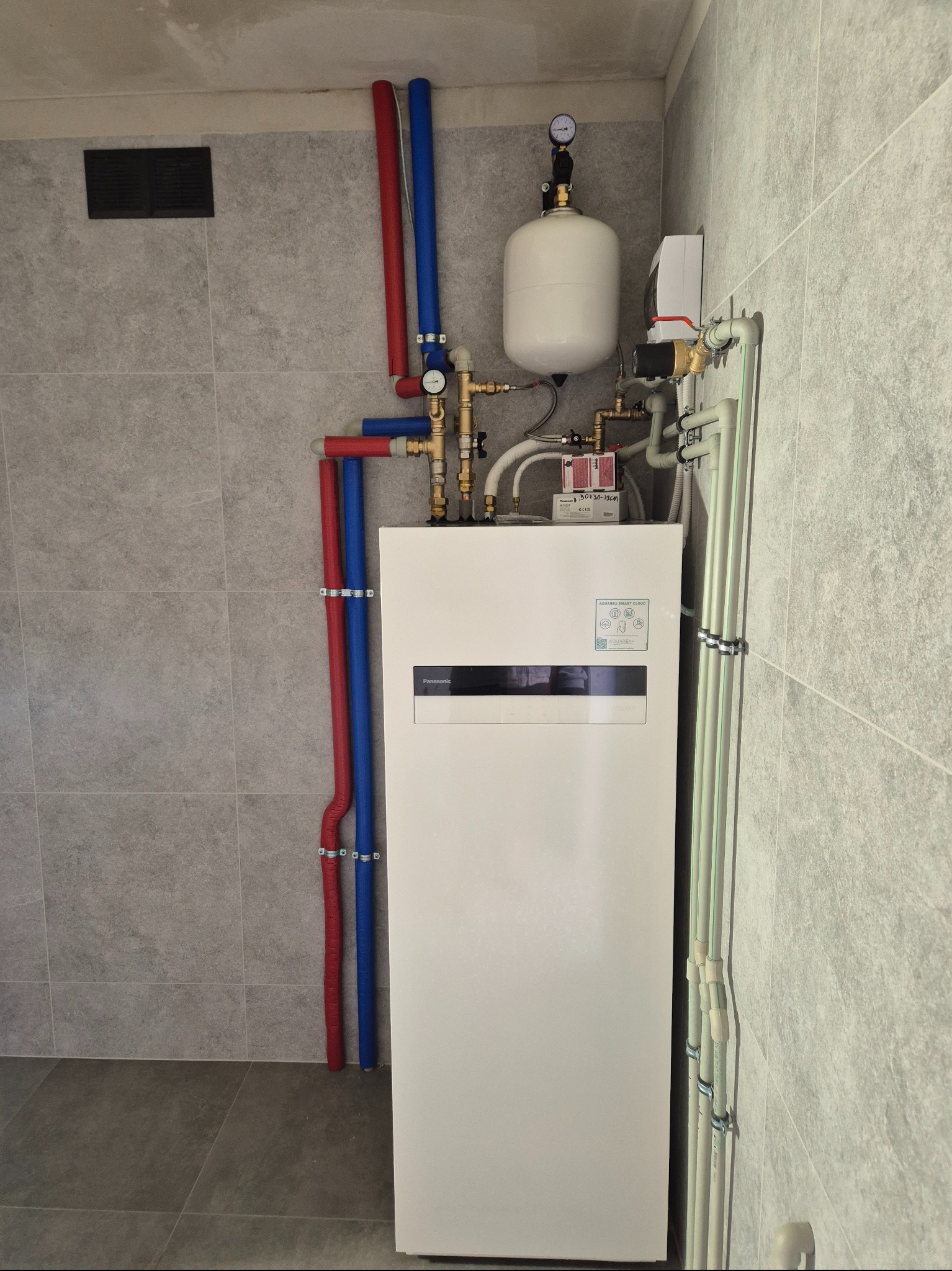Nowoczesna, kompaktowa pompa ciepła Panasonic Aquarea z podłączonymi rurami w izolacji czerwonej i niebieskiej, zainstalowana w pomieszczeniu wykończonym płytkami w odcieniach szarości.
