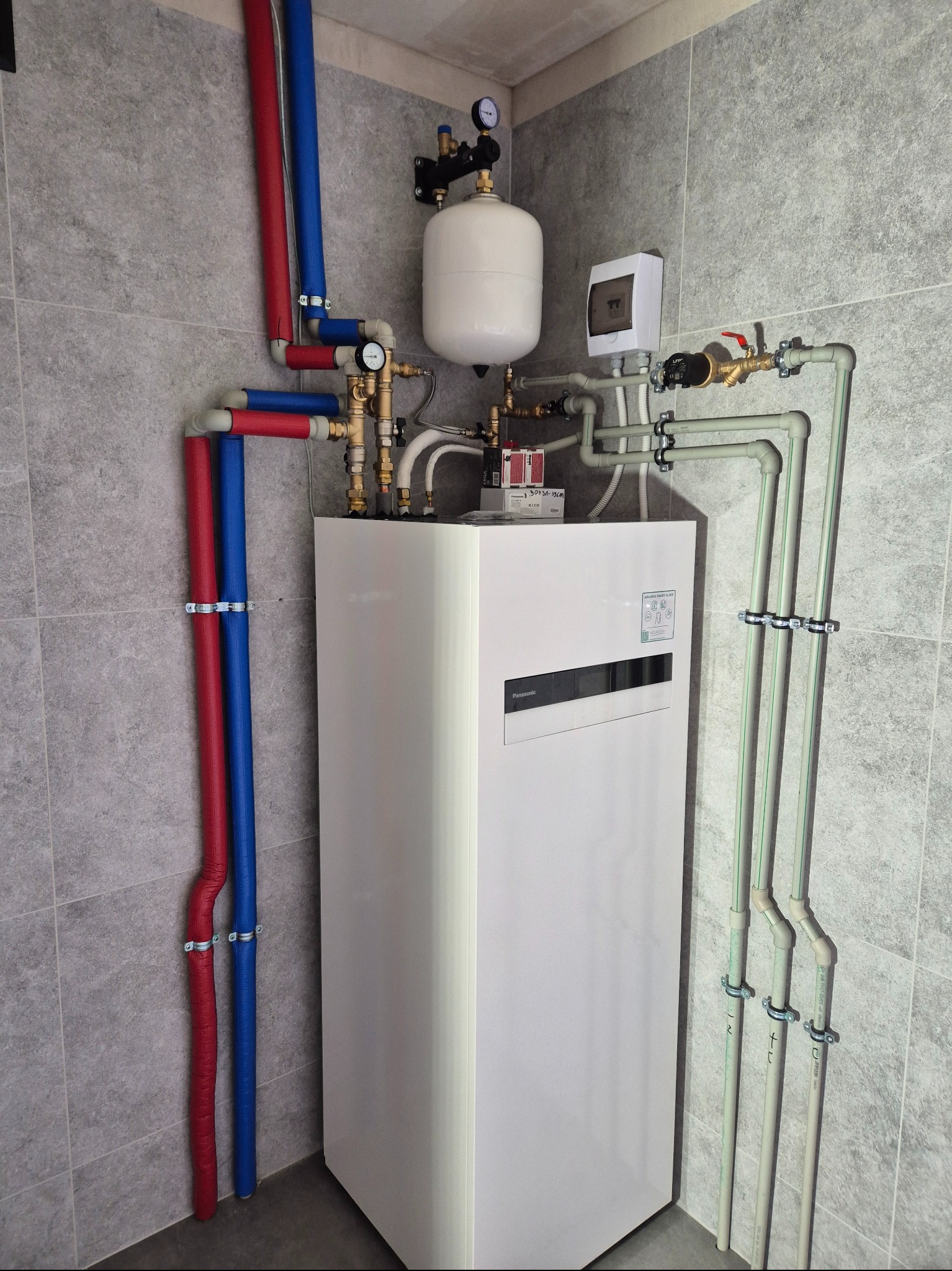 Biała jednostka wewnętrzna z podłączonymi rurami w kolorze czerwonym, niebieskim i szarym na tle szarych płytek ściennych. Widoczne manometry i zawory.