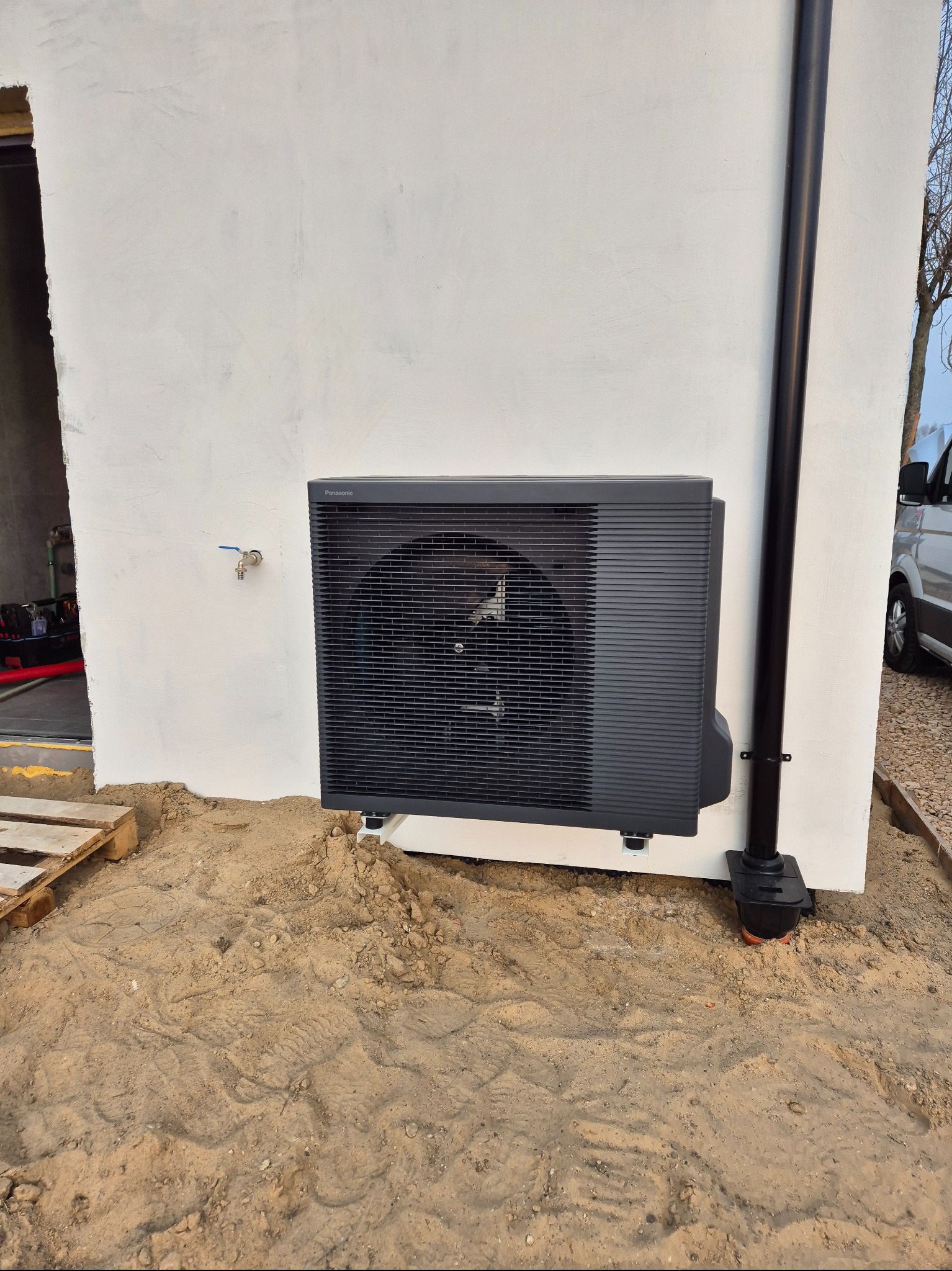 Zewnętrzna jednostka klimatyzacji Panasonic zamontowana na białej ścianie budynku, obok czarnej rury spustowej, z widocznym fragmentem zaparkowanego vana w tle, na ziemi piasek i ślady stóp.