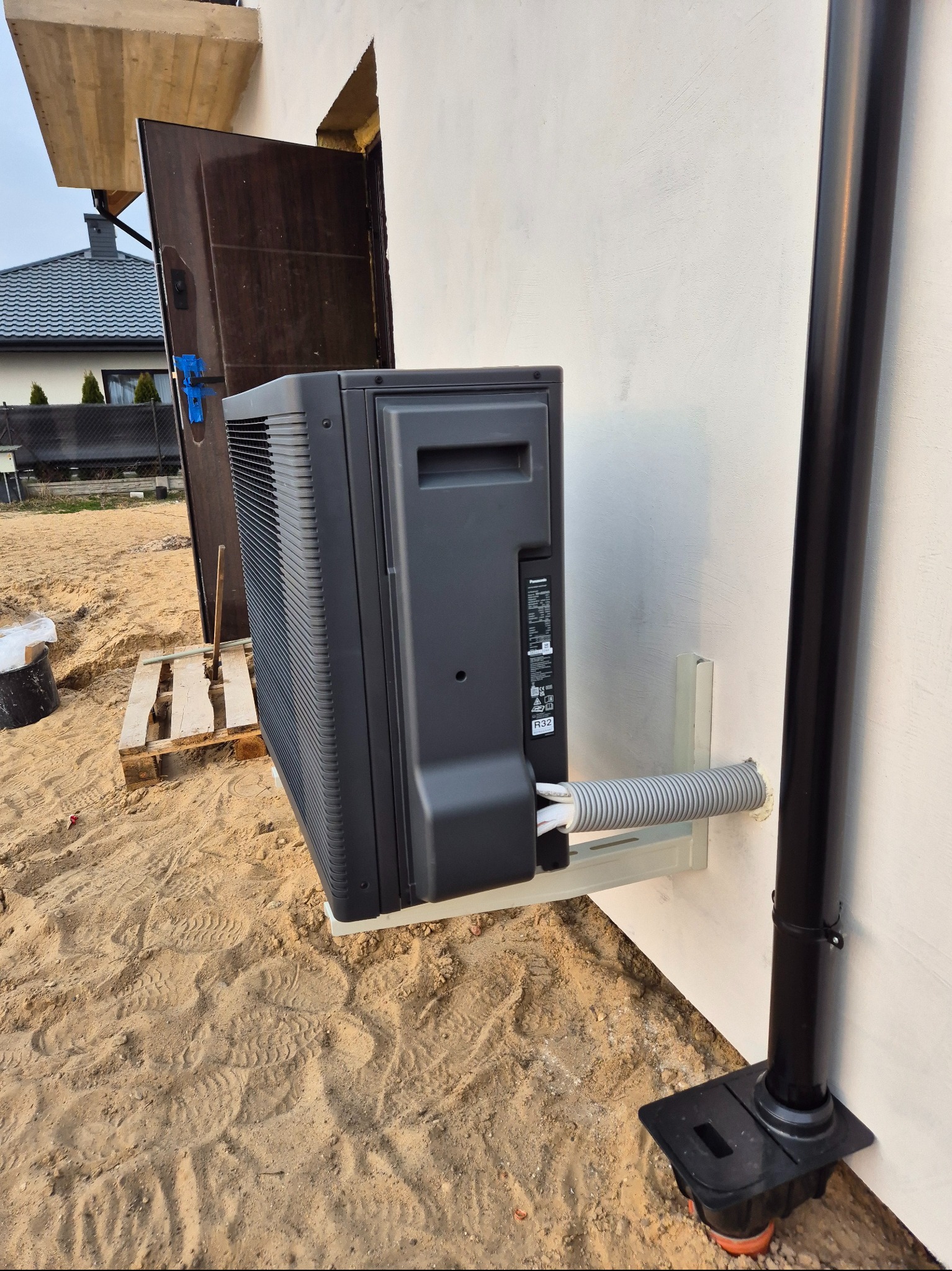 Czarna jednostka zewnętrzna pompy ciepła marki Panasonic zamontowana na wsporniku przy białej ścianie budynku w trakcie instalacji, widoczne przewody w szarej karbowanej osłonie oraz rura spustowa.