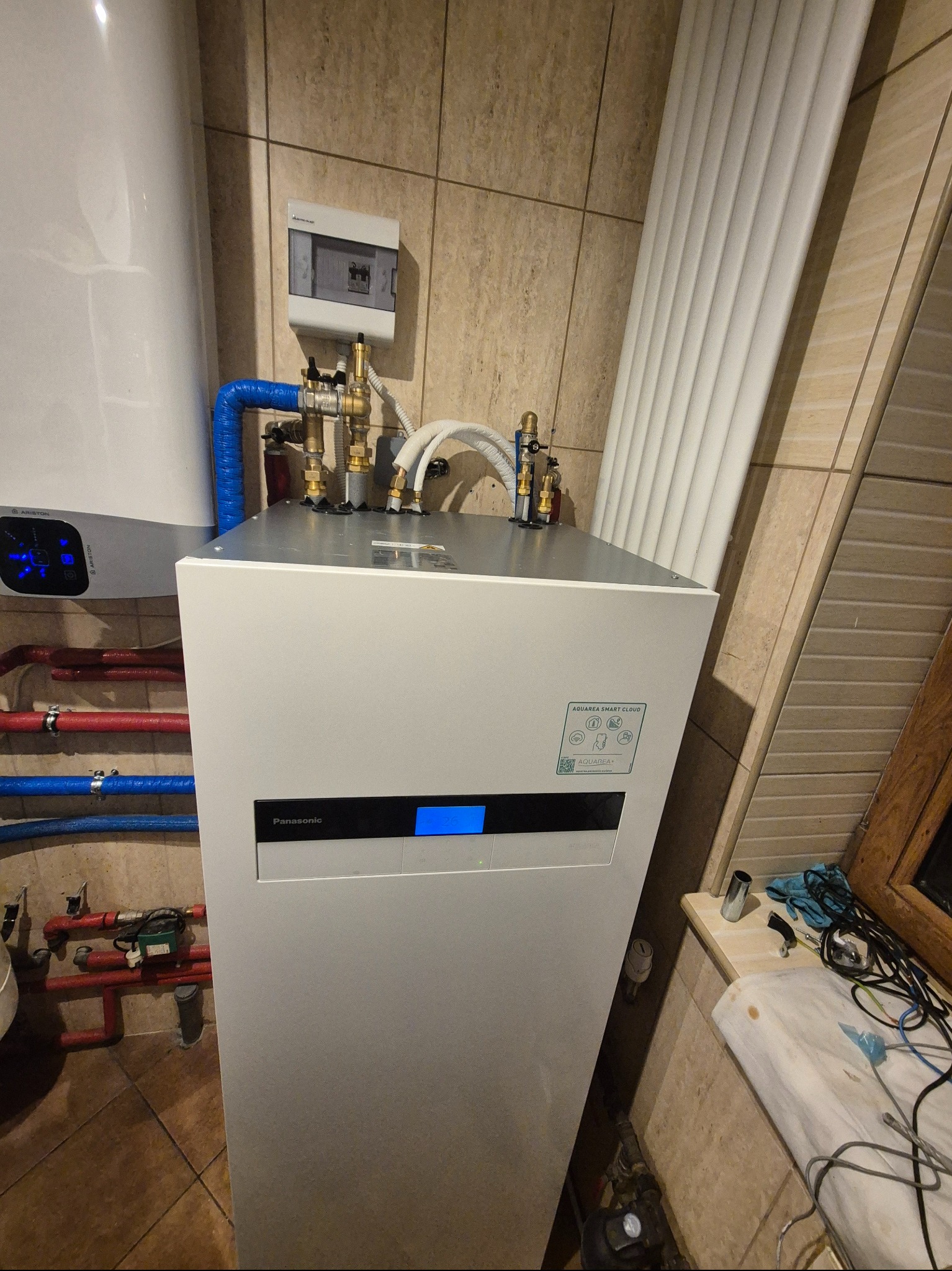 Biała pompa ciepła Panasonic Aquarea Smart Cloud z niebieskim wyświetlaczem, podłączona rurami w kolorze czerwonym i niebieskim, widoczna instalacja elektryczna i grzejnik panelowy w tle, płytki...