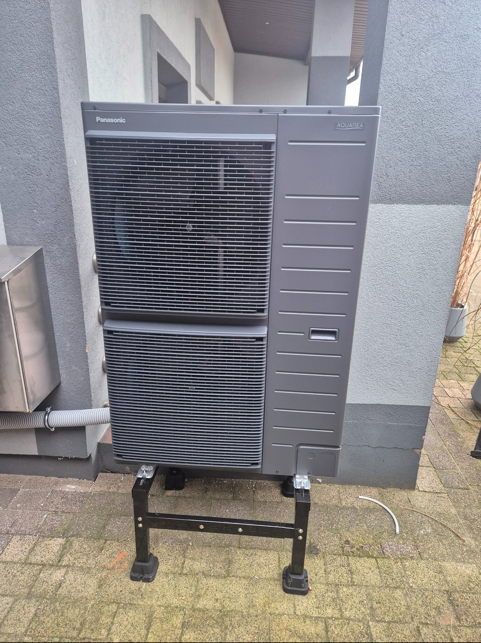 Szara pompa ciepła Panasonic Aquarea na czarnym stelażu, ustawiona na kostce brukowej przy szarej ścianie budynku z widoczną instalacją odprowadzającą skropliny.
